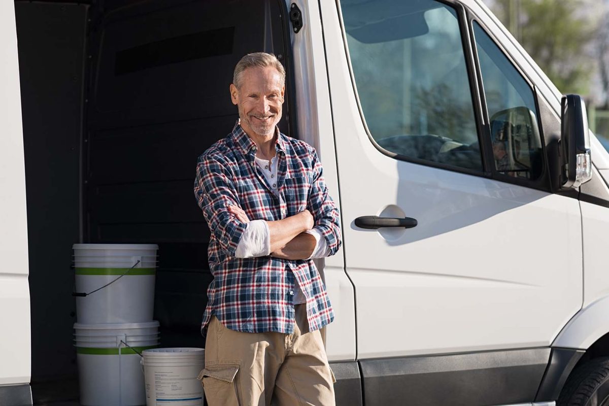 Portrait of painter standing next to his van with paint container. Decorator standing next to van and looking at camera. Happy smiling worker leaning on his white truck with folded arms.